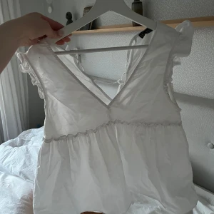 Vit blus från Zara - Säljer en söt vit blus från Zara med volangdetaljer och v-ringning. Blusen har korta ärmar och knytning i ryggen. Perfekt för en somrig look! 🌸köpt för väldigt flera år sen så ganska säker på att den inte säljs längre 