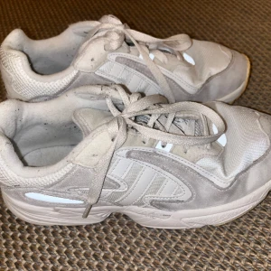 Vita sneakers från Adidas - Säljer ett par vita sneakers från Adidas med snörning och rund tå. Skorna har en stilren design med diskreta detaljer och är perfekta för vardagsbruk.