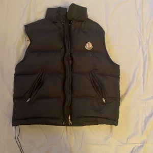Svart dunväst från Moncler - Säljer en snygg svart dunväst från Moncler med dragkedja och två fickor framtill. Västen har en hög krage och är perfekt för kyligare dagar. Insidan är beige och ger en fin kontrast. Passar bra till både casual och mer uppklädda tillfällen.