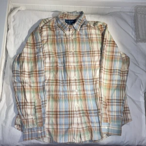 Rutig skjorta från Ralph Lauren - Säljer en stilren rutig skjorta från Ralph Lauren i mjuka toner av blått, brunt och grönt. Skjortan har långa ärmar och knappar framtill. Den är broderad med Ralph Laurens ikoniska logga på bröstet. Skjortan är i storleken XL.                              Priset är inte hugget i sten.                                        Liknande plagg finns på profilen, mvh