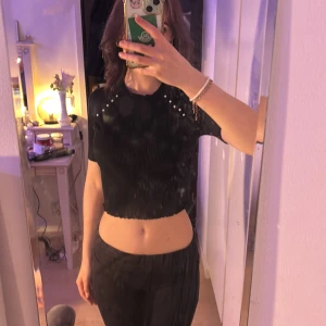 Svart ribbad croppad topp med pärldetaljer - Snygg svart ribbad topp med korta ärmar och pärldetaljer vid axlarna. Den är croppad och passar perfekt för en trendig look.