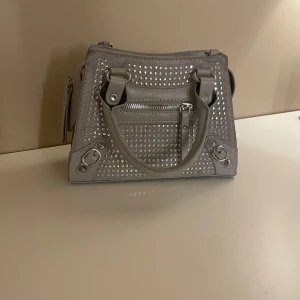 Grå handväska med nitar - Snygg grå handväska dekorerad med nitar och dragkedja framtill. Väskan har två handtag och en stilren design som passar perfekt för en trendig look. Perfekt för att ge din outfit en extra edge.
