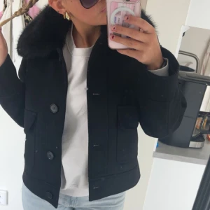 Svart jacka med pälsdetalj - Snygg svart jacka med stor krage i pälsimitation och knappar framtill. Jackan har två stora fickor med lock, jag har kvar kvittot på den och den är använd ca 5 gånger! Så den är i nyskick 