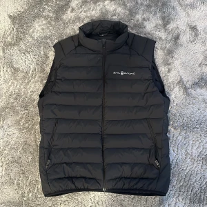 Svart dunväst från Sail Racing - Snygg svart dunväst från Sail Racing med dragkedja och hög krage. Perfekt för kyliga dagar och ger en sportig look. Två praktiska fickor med dragkedja framtill.