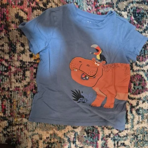 Blå t-shirt med flodhästmotiv från C&A - Säljer en blå t-shirt från C&A med ett roligt flodhästmotiv. Flodhästen har en öppningsbar mun med en bil inuti och en fågel på ryggen. Perfekt för barn som gillar djur och äventyr!