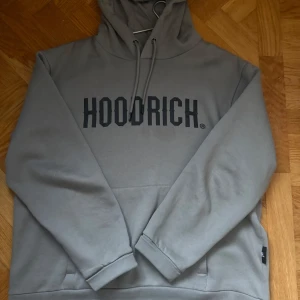 Grå hoodie från Hoodrich - Säljer en stilren grå hoodie från Hoodrich med svart text på bröstet. Tröjan har en klassisk design med huva och dragsnören samt en stor magficka. Perfekt för en avslappnad stil.