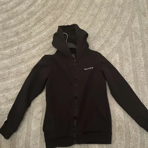 Svart hoodie från McKenzie - Säljer en svart hoodie från McKenzie med dragkedja och huva. Den har långa ärmar och en enkel design, perfekt för en avslappnad stil. Passar bra till både träning och vardag.