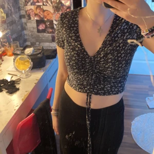 Svart blommig croppad topp - Säljer en svart croppad topp med små vita blommor. Toppen har korta ärmar och en v-ringad design med knytning framtill.