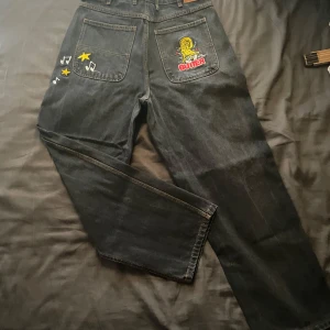 Butter Jeans - Coola svarta baggy butter jeans med unika broderier av Smurfarna'. Jeansen har en avslappnad passform och är dekorerade med stjärnor och musiknoter. Perfekta för en avslappnad stil med en twist.