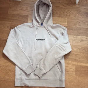 Beige hoodie från Jack & Jones - Snygg beige hoodie från Jack & Jones Originals med svart tryck på bröstet. Tröjan har huva med snörning, känguruficka och mjukt fleecefoder på insidan. Perfekt för dig som gillar en clean och stilren look. Perfekt nu framåt vintern med ett litet kallare klimat. Nypris 500