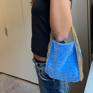 Himmelsblå virkad Stella McCartney väska med guld kedja - Superfin axelväska i ljusblått virkat material med chunky guldfärgad kedja som axelrem. Väskan är mjuk och har en cool, handgjord känsla. Perfekt storlek för det viktigaste och ger en unik touch till din outfit. 