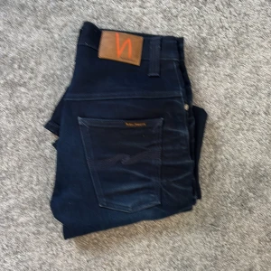 Nästan HELT NYA jeans från Nudie  - Snygga nästintill HELT NYA mörkblå jeans från Nudie Jeans Co med klassisk femficksdesign och kontrastsömmar på bakfickan. Jeansen har normal passform och är tillverkade i slitstarkt denimtyg av bomull. Perfekta för dig som gillar stilrena och tidlösa jeans.