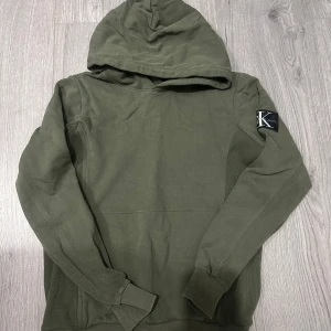 Olivgrön hoodie från Calvin Klein Jeans - Snygg olivgrön hoodie från Calvin Klein Jeans med stor huva och ribbade muddar. Klassisk logga på vänster ärm som ger en cool touch. Perfekt för dig som gillar stilrena och bekväma plagg med streetkänsla.