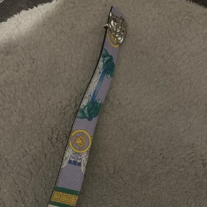 Färgglatt Versace mönstrat bälte med silverdetalj - Unikt bälte med färgstarkt mönster i lila, grönt, gult och blått. Bältet har en cool silverfärgad spänne med lejonhuvud och detaljerade ornament. Perfekt för att lyfta vilken outfit som helst med en touch av lyxig vibe.