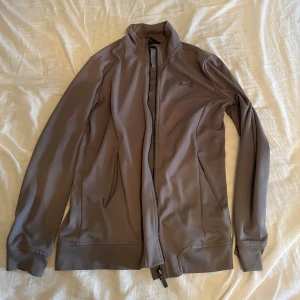 Beige vindjacka från A–Z, stl M - Säljer en stilren tröja från A–Z i storlek M. Tröjan har hel dragkedja framtill, höga muddar och två fickor med dragkedja. Materialet är mjukt och stretchigt, perfekt för en aktiv vardag. Enkel design med A–Z-logga på bröstet.