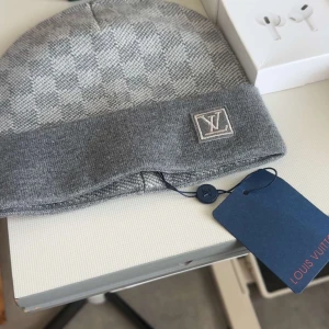 Grå rutig mössa från Louis Vuitton - Exklusiv grå mössa från Louis Vuitton med klassiskt rutigt mönster och vikt kant. LV-logga framtill och snyggt vävd struktur. Perfekt accessoar för dig som vill ha en stilren och lyxig look under kalla dagar.