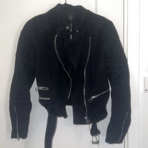 Svart mockajacka med bälte och dragkedjor - Snygg svart mockajacka från Outerwear med coola silverfärgade dragkedjor, bälte i midjan och flera fickor. Jackan har en klassisk bikerstil och långa ärmar med dragkedjor vid ärmsluten. Perfekt för dig som vill ha en edgy look.