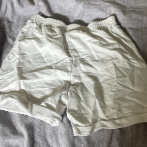 Vita shorts med blå text 'Lovely girl' - Vita shorts i mjuk bomull med resår i midjan och avslappnad passform. På baksidan finns trycket 'Lovely girl' i blå gotisk stil. Perfekta för chill dagar eller när du vill ha en enkel och snygg look.