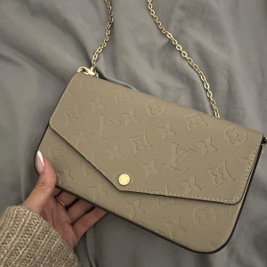 Beige axelremsväska i lyxig stil - Säljer denna supersnygga axelväska i beige med elegant präglat mönster och guldfärgad kedjerem. Väskan har kuvertdesign med tryckknapp, och är tillverkad i ett strukturmönstrat material som ger en exklusiv känsla. Plånbok ingår, se sista bild. Aldrig använd! 