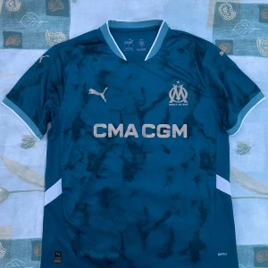 Blå Marseille fotbollströja Puma - Snygg blå Olympique de Marseille fotbollströja från Puma med klubbmärke och sponsortryck på bröstet. Tröjan har korta ärmar, ljusblå detaljer vid krage och ärmslut samt ett unikt marmorerat mönster. Tillverkad i lätt och ventilerande material.