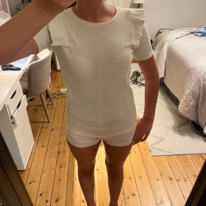 Vit ribbad topp med volangärm - Snygg vit ribbad topp med korta ärmar och volangdetaljer på axlarna. Perfekt för dig som gillar en enkel men ändå trendig look. Rund halsringning och figurnära passform gör den lätt att matcha med allt.