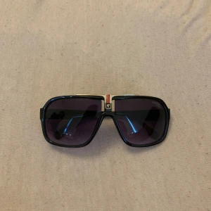 Svarta Carrera solglasögon med guld - Snygga svarta Carrera solglasögon med fyrkantig form och mörka linser. Bågarna har guldiga detaljer och Carrera-logga på sidorna. Framtill finns en cool detalj i guld och rött ovanför näsryggen. Perfekta för dig som vill sticka ut med en retrovibe.