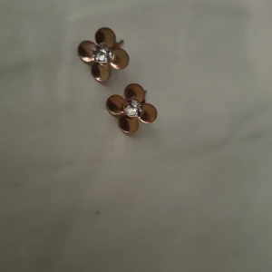 Guldiga blomformade örhängen - Säljer ett par söta örhängen i form av blommor med fyra blad i guldig metall och en glittrande sten i mitten. Perfekta för dig som gillar en feminin och stilren look med lite extra bling.