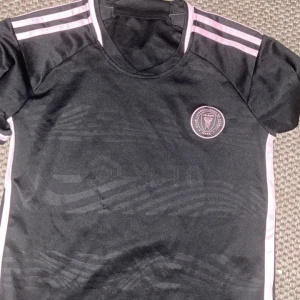 Inter Miami Messi 10 matchtröja - Snygg svart Inter Miami fotbollströja från Adidas med rosa detaljer och Messi 10-tryck på ryggen. Tröjan har korta ärmar, rund hals och klubbmärke på bröstet. Tillverkad i lätt och ventilerande material, perfekt för fotbollsplanen eller att bära som supporter.