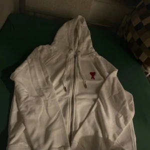 Vit hoodie med rött hjärta från Ami - Vit hoodie från Ami Alexandre Mattiussi med dragkedja, huva och snörning. På bröstet finns ett broderat rött hjärta med bokstaven A. Materialet är mjuk bomull och ryggen har diskret AMI-broderi. Perfekt för en clean och stilren look.