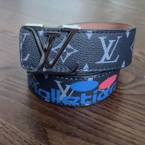 Svart bälte - Snyggt svart bälte med klassiskt monogrammönster och stora spännet i silver. Bältet har detaljer i blått, vitt och gult samt inslag av olika tryck och patchar. Insidan är brun och materialet är skinn.