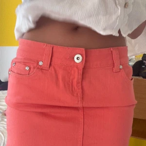 Korallrosa jeanskjol med strassdetaljer - Säljer en snygg korallrosa kort jeanskjol med fem fickor, bälteshällor och silverfärgade nitar. Baksidan har en cool strassdekor på ena fickan. Kjolen har klassisk knapp och dragkedja framtill. Perfekt för dig som vill sticka ut lite extra!