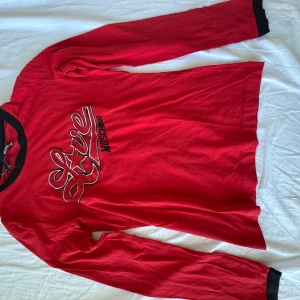 Röd långärmad tröja Love Moschino - Röd långärmad tröja från Love Moschino med svart krage och svarta. Framsidan har ett stort broderat Love Moschino-tryck i röd, svart och vitt. Tröjan är i mjuk bomull och har en klassisk, normal passform.