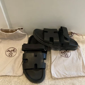 Svarta sandaler  - Svarta sandaler i stilren design med breda remmar och klassiskt H-utskuret mönster. Tillverkade i skinn med justerbar kardborre upptill för extra komfort. Perfekta för en avslappnad och lyxig look.