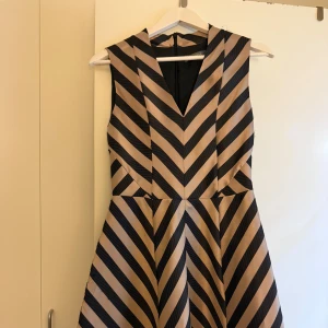 Randig kortklänning från Oasis - Supersnygg ärmlös kortklänning från Oasis med svart och beigea diagonala ränder. Klänningen har v-ringning, markerad midja och dragkedja bak. Perfekt för dig som gillar grafiska mönster och vill sticka ut lite extra.