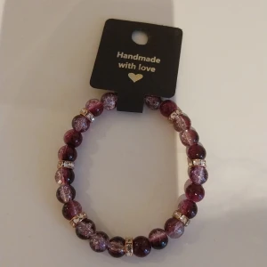 Lila pärlarmband med strassdetaljer - Handgjort armband med lila och vinröda pärlor, dekorerat med glittrande strassringar mellan pärlorna. Elastiskt band gör det enkelt att ta på och av. Perfekt accessoar för att lyfta din outfit med lite extra bling.