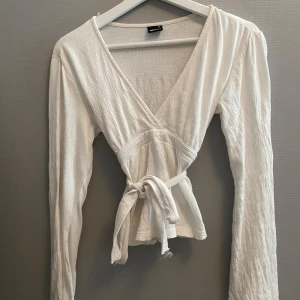 Vit omlottopp med knyt från Gina Tricot - Supersöt vit omlottopp från Gina Tricot med långa ärmar och knyt i midjan. Toppen har v-ringning och är lätt croppad, vilket ger en fräsch och trendig look. Tillverkad i ett mjukt, strukturerat bomullstyg som känns skönt mot huden.