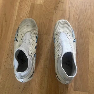 Nike vita fotbollsskor med blå detaljer elite - Säljer ett par vita Nike fotbollsskor med blå och svarta detaljer samt rosa inslag på sulan. Skorna har en strumpliknande passform och är tillverkade i syntetmaterial för bästa grepp och känsla på planen. Perfekta för dig som vill sticka ut med stilrena och sportiga skor.