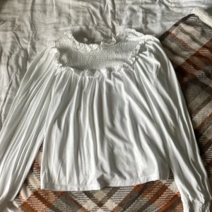 Vit långärmad blus med smock - Säljer en söt vit blus med lång ärm och smockad överdel. Blusen har volangdetaljer vid halsringningen och är tillverkad i ett mjukt, lätt tyg som faller snyggt. Perfekt för dig som gillar romantisk och luftig stil.