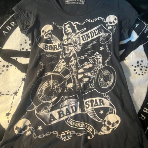 Svart t-shirt med tryck från Liquorbrand - Cool svart t-shirt från Liquorbrand med stort rockigt tryck framtill. Motivet har en tatuerad kvinna, motorcykel, dödskallar och kedjor samt texten 'Born Under A Bad Star'. Klassisk passform och rund hals.
