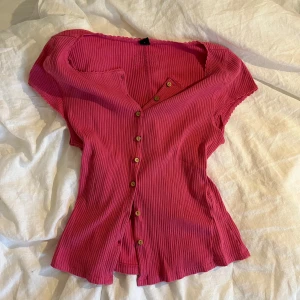 Rosa ribbad blus med knappar - Supersöt rosa ribbad blus med korta ärmar och träknappar hela vägen framtill. Blusen har v-ringning och små volangdetaljer vid ärmslut och halsringning. Perfekt för dig som gillar färg och vill ha en feminin touch på outfiten. OBS! Kan skicka som tidigast 8e augusti