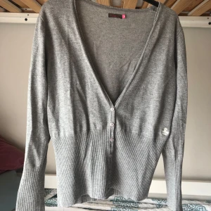 Grå cardigan från Hillevi med knappar - Grå cardigan/kofta från Hillevi med djup v-ringning och knappar framtill. Tröjan har långa ärmar och ribbad nederdel samt muddar. Liten broderad krona på ena sidan. Väldigt stretchigt och mjukt material.