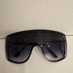 Svarta shield-solglasögon - Stora, futuristiska shield-solglasögon med svart båge och mörka, tonade linser. Rektangulär form som täcker mycket av ansiktet och ger en riktigt cool vibe. Perfekta för dig som vill sticka ut och skydda ögonen mot solen.