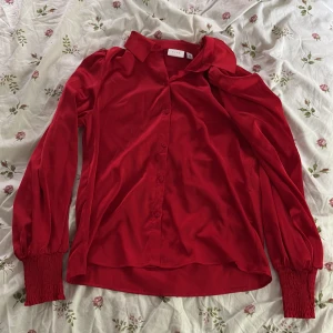 Röd skjorta med puffärm från Vila - Snygg röd skjorta från Vila i storlek 34. Skjortan har långa puffärmar med breda smockade muddar, klassisk krage och knäppning framtill. Materialet är glansigt och mjukt, vilket ger en lyxig känsla. Perfekt för dig som vill sticka ut med färg.