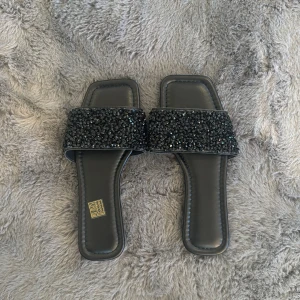 Svarta sandaler med glittriga detaljer - Snygga svarta sandaler med fyrkantig tå och bred rem täckt av svarta glittriga stenar. Perfekta för att lyfta vilken outfit som helst med lite extra bling. Platt sula och stilren design gör dem både trendiga och bekväma.