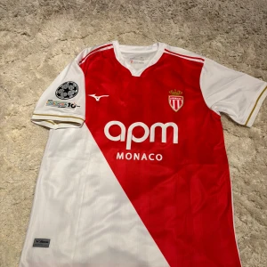 AS Monaco Pogba 8 matchtröja Mizuno L - AS Monaco fotbollströja med Pogba 8 på ryggen. Klassisk röd och vit design med diagonal färgdelning, klubbmärke och sponsortryck framtill. Kortärmad modell i lätt och ventilerande material, officiell Champions League-patch på ärmen. Perfekt för dig som älskar fotboll och stil.