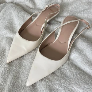 Vita spetsiga ballerinaskor från Mango - Snygga vita ballerinaskor från mango med spetsig tå och smal rem runt hälen. Skorna har en stilren design och är gjorda i syntetmaterial med slät yta. Perfekta för dig som gillar minimalistisk och chic stil.