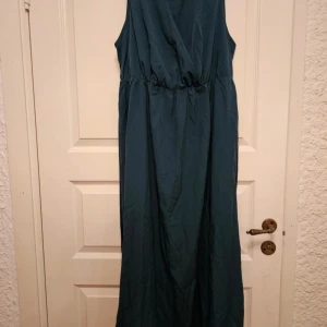 Mörkgrön långklänning från Seasty Curve - Snygg mörkgrön långklänning från Seasty Curve i storlek 2XL. Klänningen är ärmlös med omlottliv och har ett matchande bälte i midjan. Perfekt för dig som gillar en enkel men elegant look.