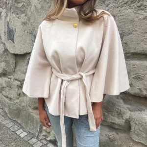 Beige poncho - Stilren beige poncho med knytbälte i midjan. Hög krage med dekorativ guldfärgad knapp upptill. Perfekt för dig som vill ha en chic och modern look till vardags. ENDAST ANVÄND TVÅ GÅNGER!