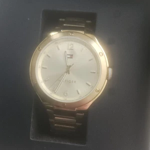 Guldfärgad klocka Tommy Hilfiger - Stilren guldfärgad klocka från Tommy Hilfiger med metallarmband och rund urtavla i silver. Klockan har minimalistisk design med diskreta index och Hilfiger-loggan på urtavlan. Kvitto och garanti finns!
