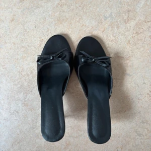 Svarat kittenheels - Stilrena svarta sandaler med låg klack och öppen tå. Skorna har en söt rosettdetalj framtill och är tillverkade i ett slätt material som ger en elegant look. Perfekta för dig som gillar en klassisk och chic vibe.